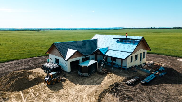 House under construction in a wide rural field, with a metal roof being installed and a worker in an orange shirt on the roof; construction equipment and materials nearby.
