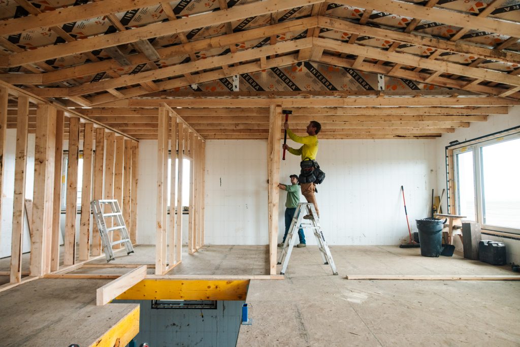 Construction workers install wooden framing inside a partially finished room; roof trusses are exposed above.